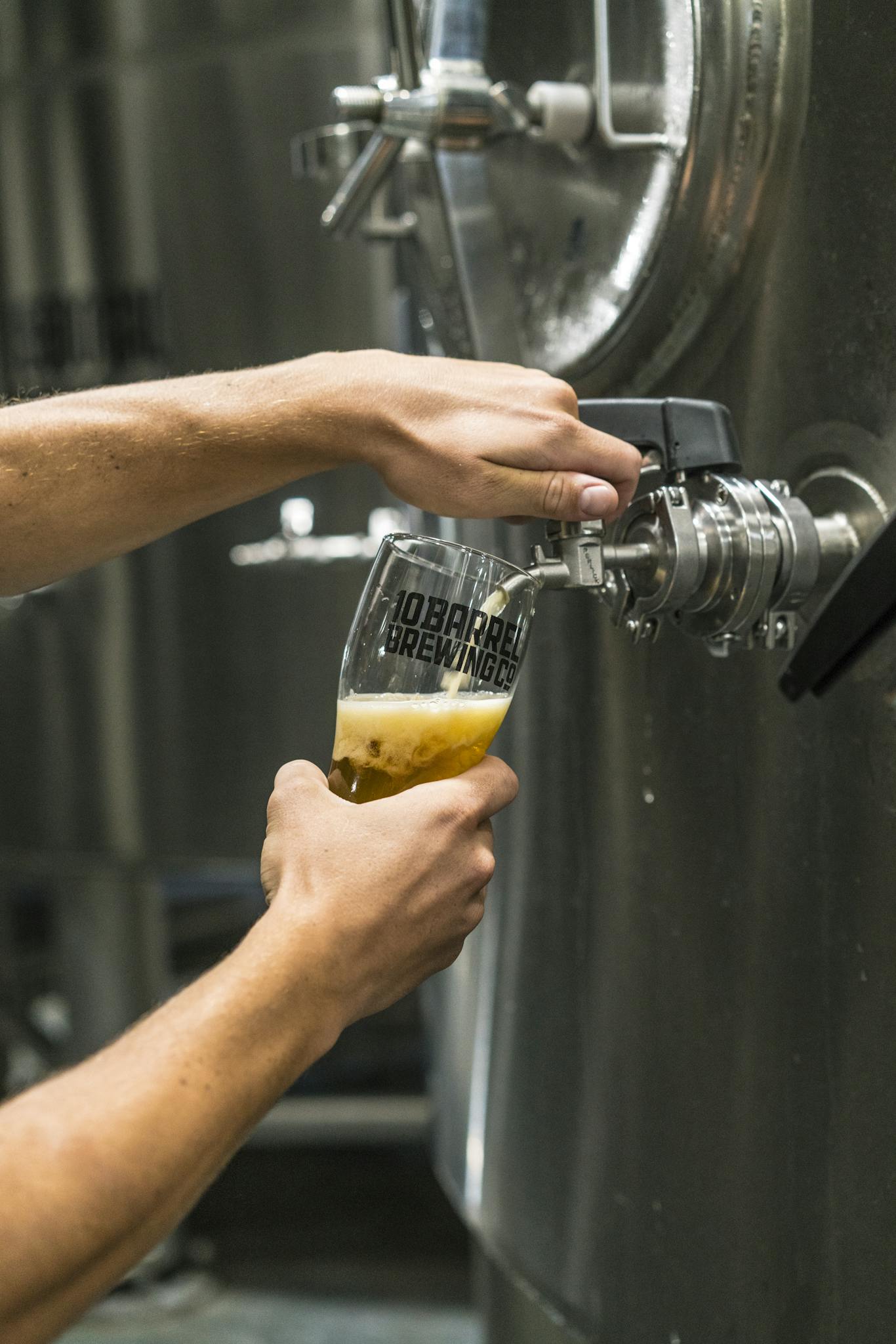 Close-up of beer being poured into a glass from a brewery tank by a person at a brewing facility.